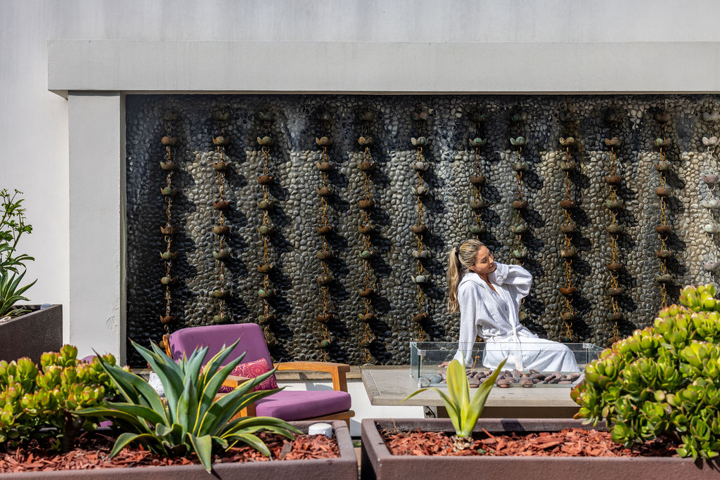 A person in a white robe sits next to a firepit in a modern outdoor patio area, with plants and a stone water feature in the background.