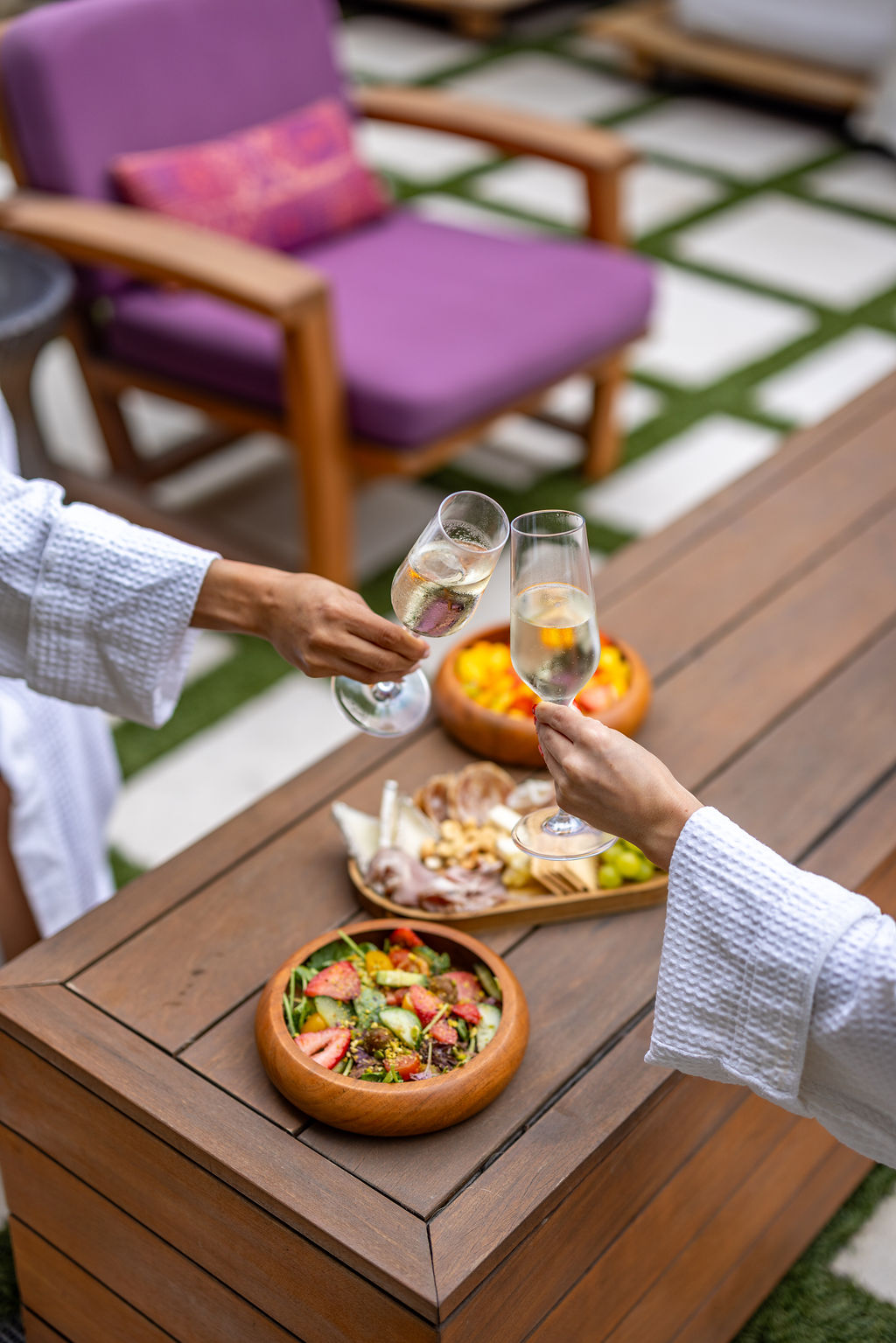 Two people in white robes clink champagne glasses over a wooden table with a salad, appetizer platter, and bowls of food; purple outdoor chair in the background.