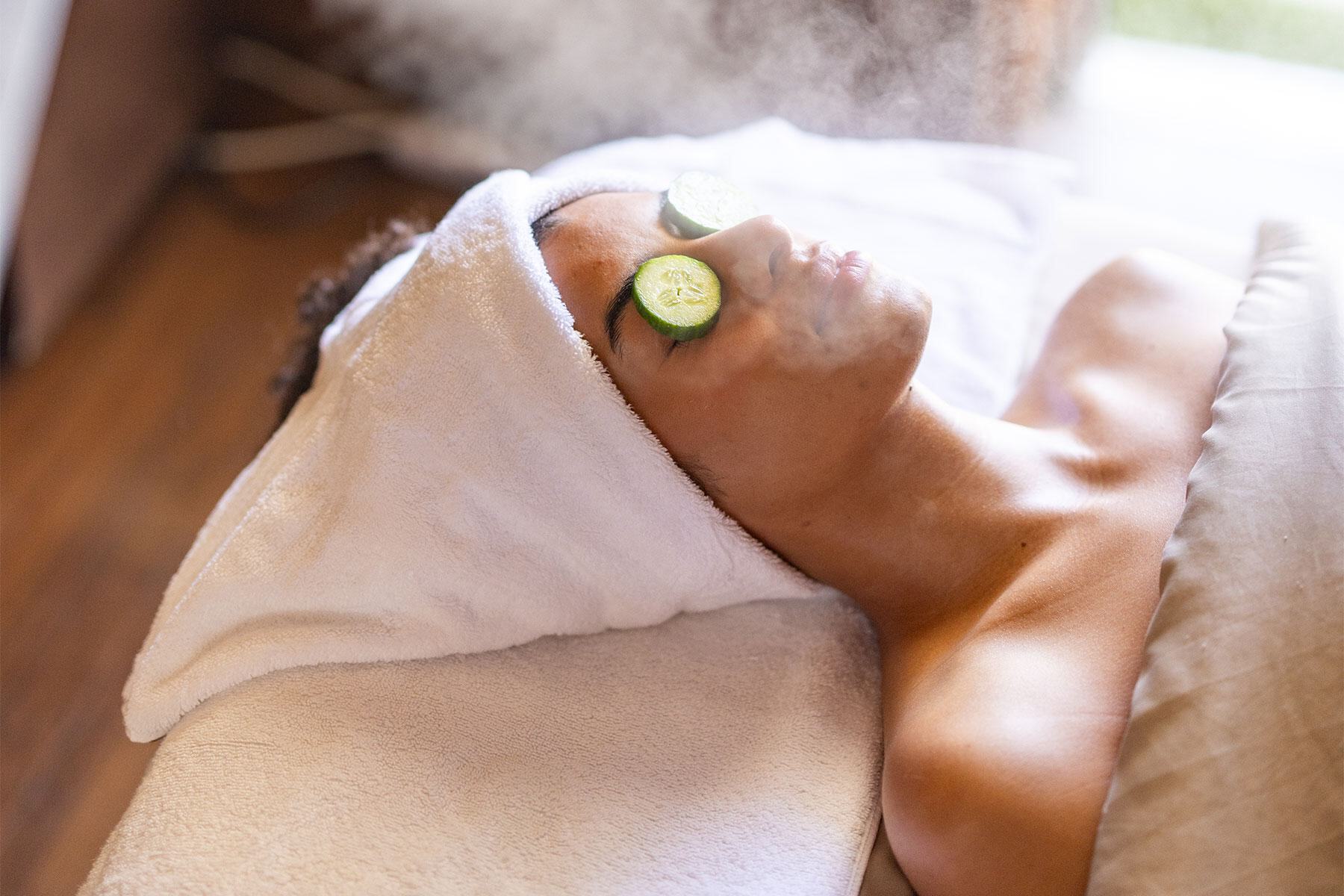 Person lying on a spa bed with a towel wrapped around their head, steam rising, and cucumber slices over their eyes—enjoying total relaxation with an Oasis Membership.