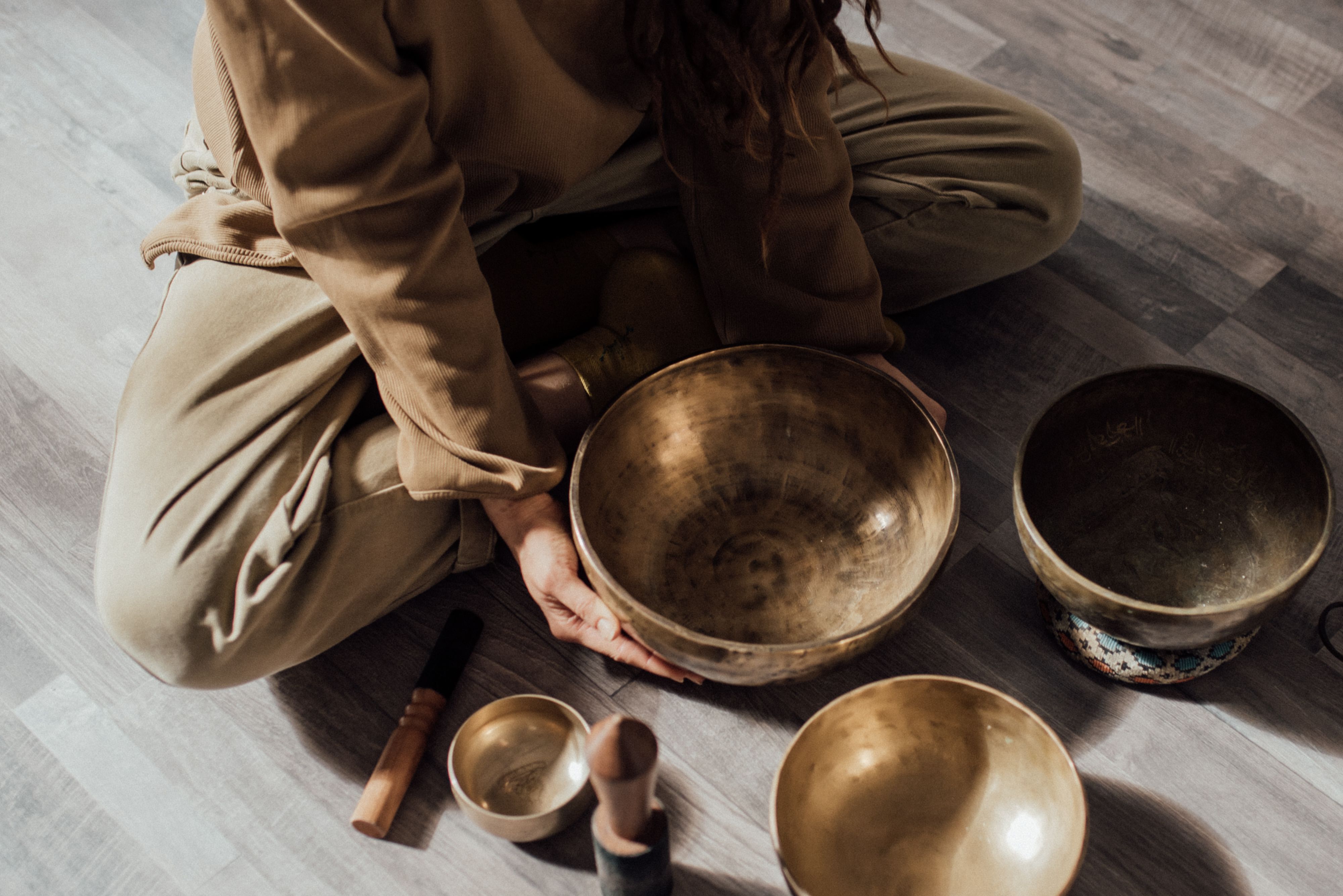 Person sitting cross-legged on the floor holding a large singing bowl, surrounded by other bowls and mallets on a wooden surface, preparing for a restorative sound bath experience—a radiant reset for mind and body.