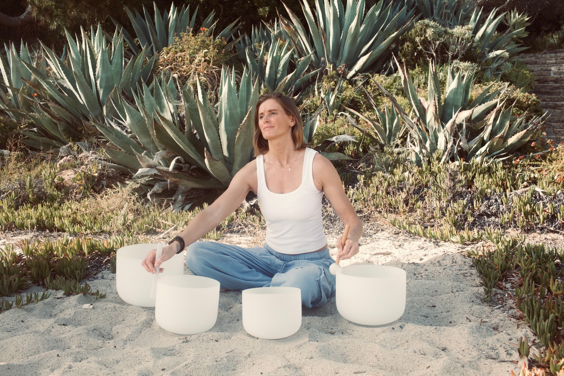 A woman sits cross-legged on sand outdoors, playing white crystal singing bowls under the Full Moon, surrounded by agave plants—a serene Radiant Reset.