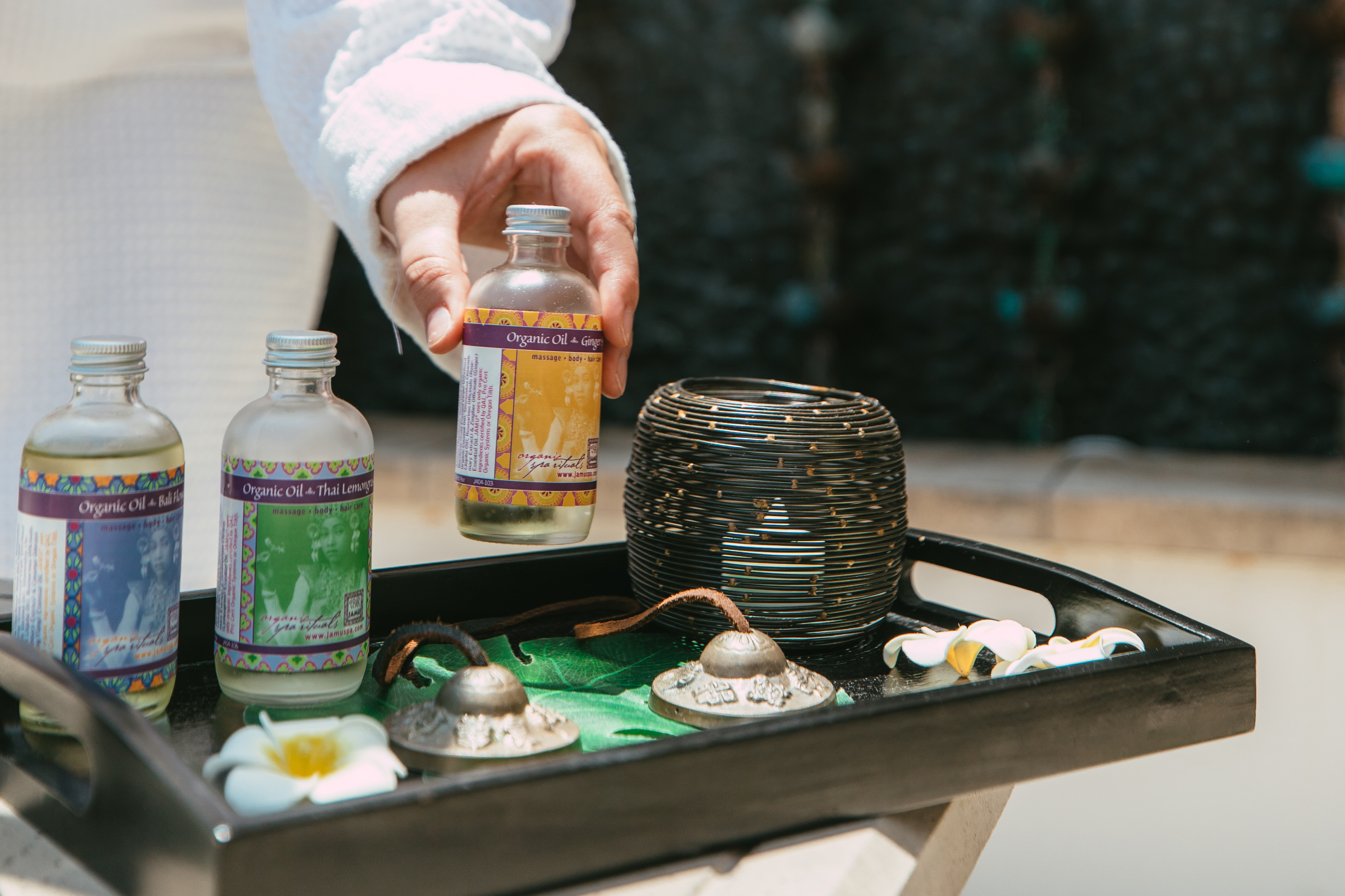 A person in a white robe holds a tray with three bottles of essential oils, a decorative candle holder, small bells, and white flowers—perfect for an Enhancement or Add-On to your relaxing spa experience.