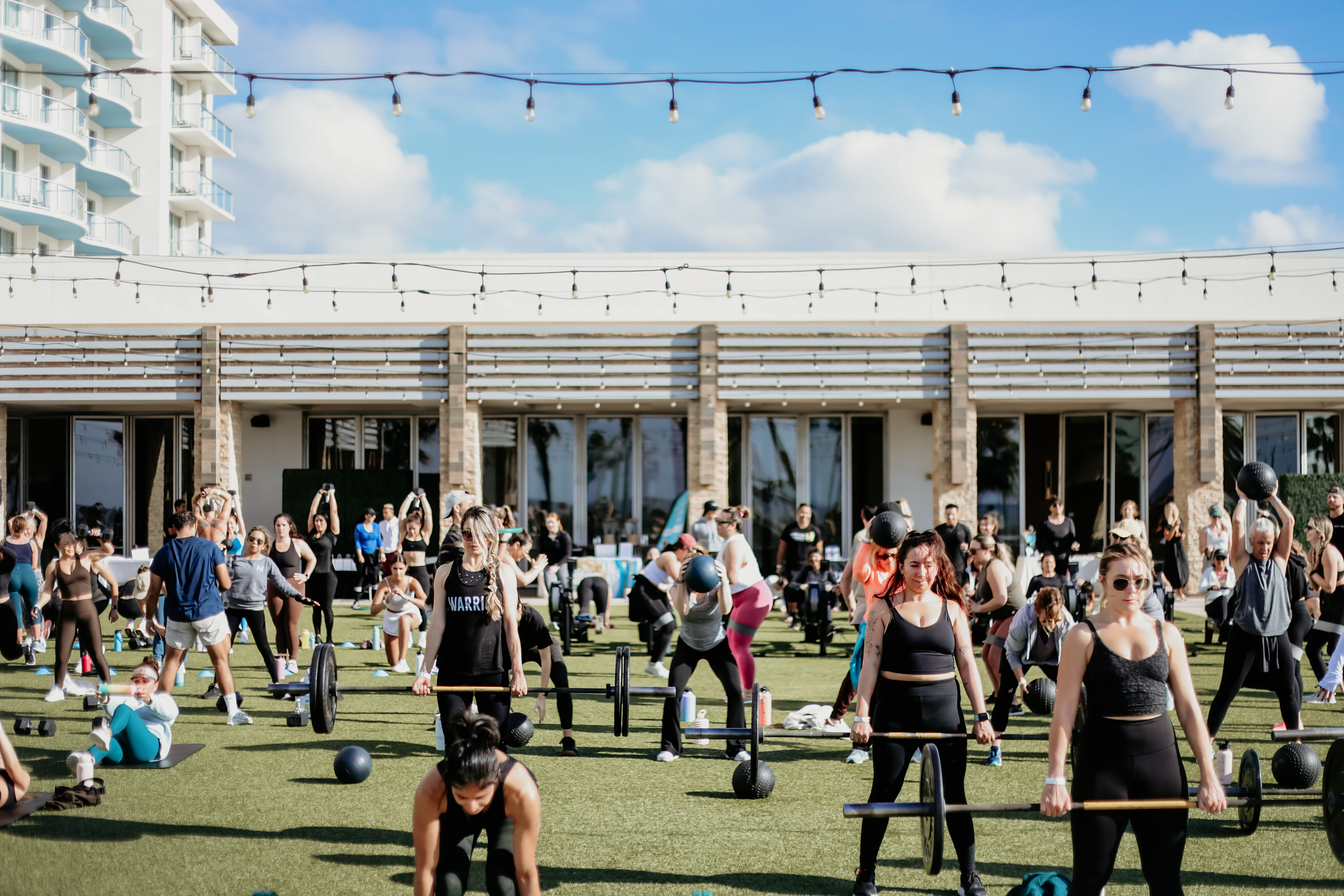 A large group of people participate in an outdoor fitness class—Waves of Wellness—involving barbells and medicine balls on a grassy area in front of a building.